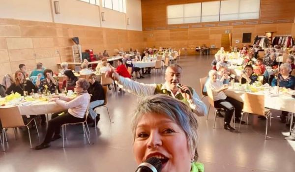 Eine Frau singt in ein Mikrofon. Dahinter steht ein Mann der ebenfalls in ein Mikrofon singt. Im Hindergrund sitzen Menschen an Tischen und klatschen.