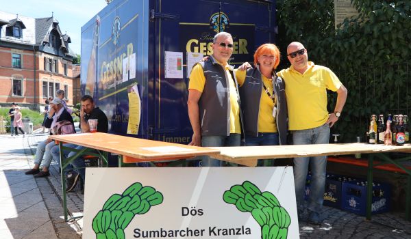 Drei Personen stehen hinter einem Verkaufsstand. Davor steht ein Schild mit der Aufschrift: Dös Sumbarcher Kranzla e.V..