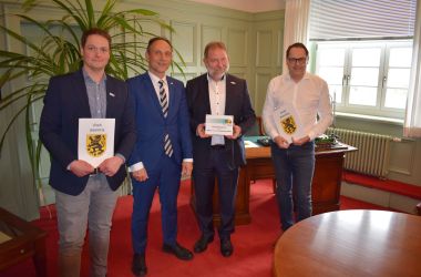 Vier Männer stehen in einer Reihe nebeneinander. Zwei von ihnen halten eine Urkunde. Ein Mann hat ein Schild zur MINT Kooperation.