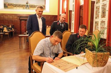 Ein Mann sitzt an einem Tisch und unterschreibt eine Urkunde. Rechts daneben sitzt ein Mann. Hinter den beiden stehen zwei weitere Männer.