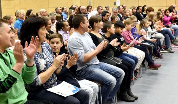 Kinder und Erwachsene sitzen in Stuhlreihen und klatschen.