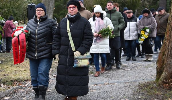 Zwei dunkel gekleidete Frauen tragen Blumengebinde zu einem Gedenkstein.