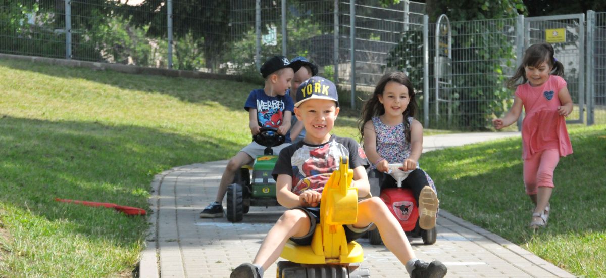 Zwei Kinder fahren auf Bobby-Car, Bagger und Taktor einen Weg entlang.