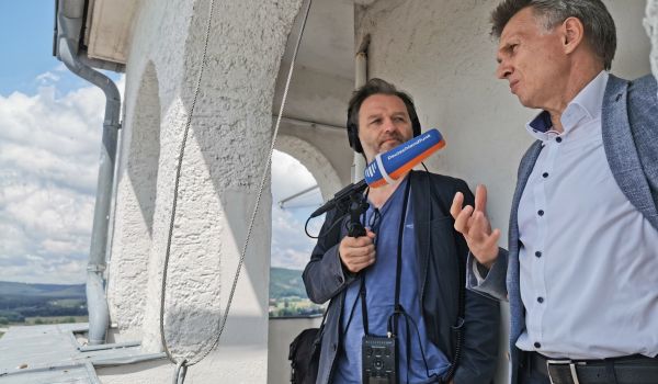 Zwei Männer stehen nebeneinander auf dem Rathausturm. Der Mann links hält eine Mikrofon in der Hand. Der Mann rechts ist der Bürgermeister Dr. Heiko Voigt.