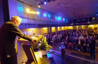 Ein Mann steht an einem Rednerpult auf einer Bühne. Im Publikum sitzen viele Menschen.