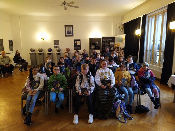 Kinder sitzen in Stuhlreihen hintereinander in einem Raum. Hinter ihnen an der Wand stehen Modelle verschiedener Planeten.