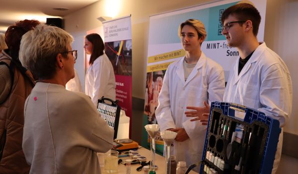 Zwei Jugendliche in weißen Kitteln erklären Experimente für Interessierte.