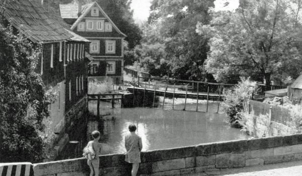 Ein Schwarz-Weiß-Foto zeigt zwei Personen, die an einer Brücke stehen und auf einen Fluss schauen. Links stehen alte Häuser.