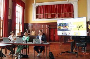 Drei Personen sitzen an Tischen nebeneinander. Auf den Tischen stehen Laptops. Daneben ist ein großer Bildschirm, der die neue Webseite der Stadt Sonneberg zeigt.