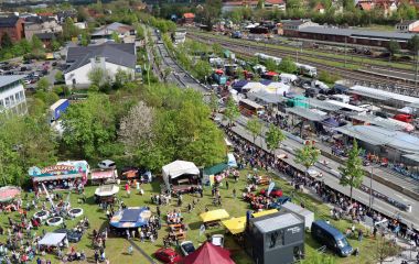 Drohnenaufnahme mit Blick auf eine Wiese und daneben das Bahnhofsgelände. Auf der Wiese sind viele Menschen und Fahrgeschäfte. Ein Teil der dazwischen liegenden Straße wurde als Rennstrecke aufgebaut.