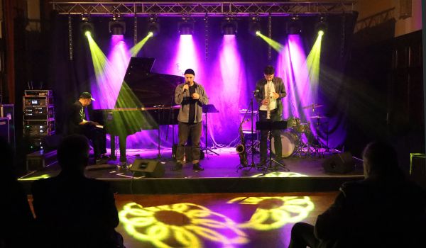 Eine Band tritt auf einer Bühne auf. Es ist dunkel. Bunte Scheinwerfer erhellen das Bild.