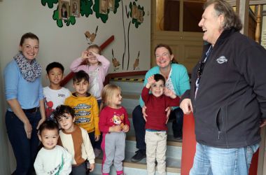 Kinder und Erwachsene stehen in einem Treppenaufgang. Im Hintergrund ist eine mit Bäumen bemalte Wand.