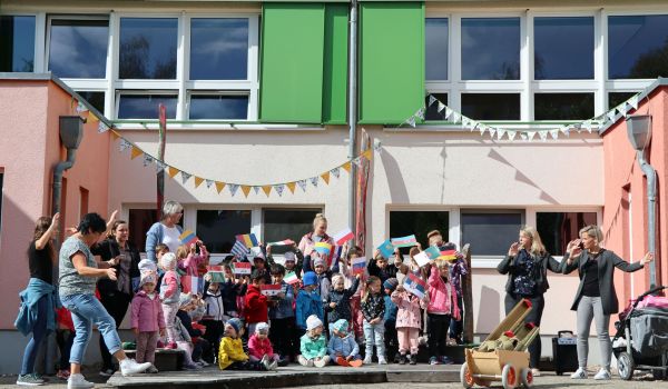 Kinder und Erwachsene stehen vor einem bunten Gebäude und halten Flaggen auf verschiedenen Ländern in den Händen.