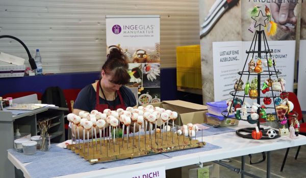Eine Frau sitzt hinter einem Tisch. Auf dem Tisch befindet sich eine Vorrichtung auf welche Glaskugeln gespießt sind. Vorne am Tisch hängt ein Schild mit der Aufschrift: Wir suchen Dich! Ausbildung 2022.