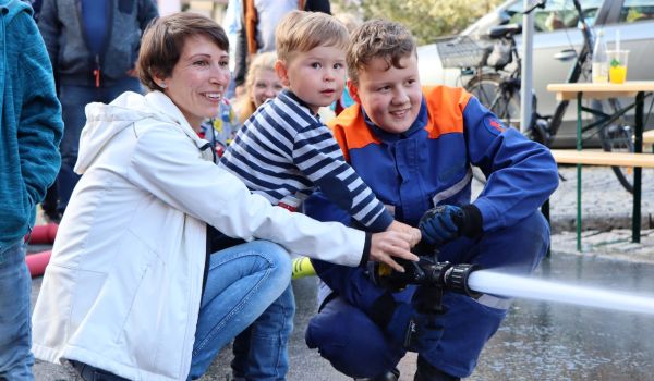 Ein junger Feuerwehrmann und eine Mutter mit Kind halten einen Feuerwehrschlauch.