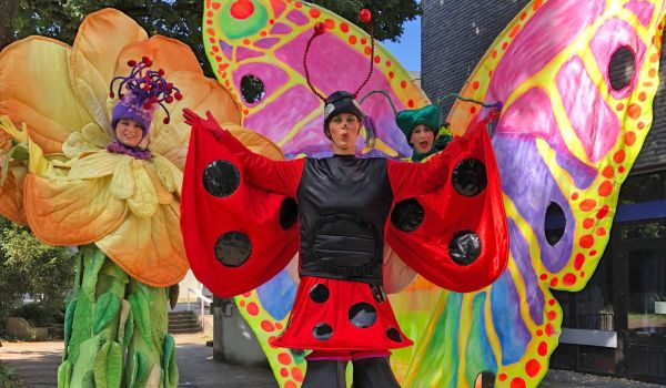 Drei Personen laufen auf stelzen und tragen bunte Kostüme. Ein Schmetterling, ein Marienkäfer und eine gelbe Blume.