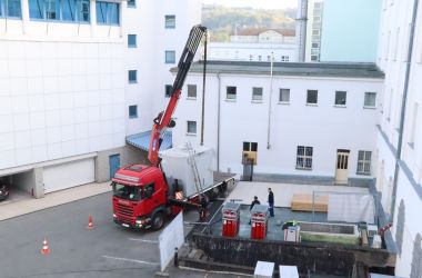 Ein roter Kran hievt einen grauen Kasten auf einen Lkw.