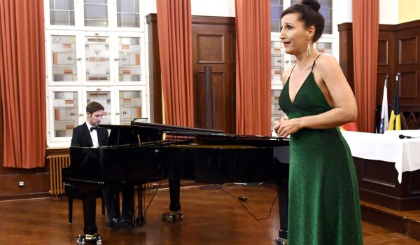 Ein Mann sitzt an einem Flügel und spielt. Davor steht eine Frau in grünem Abendkleid.