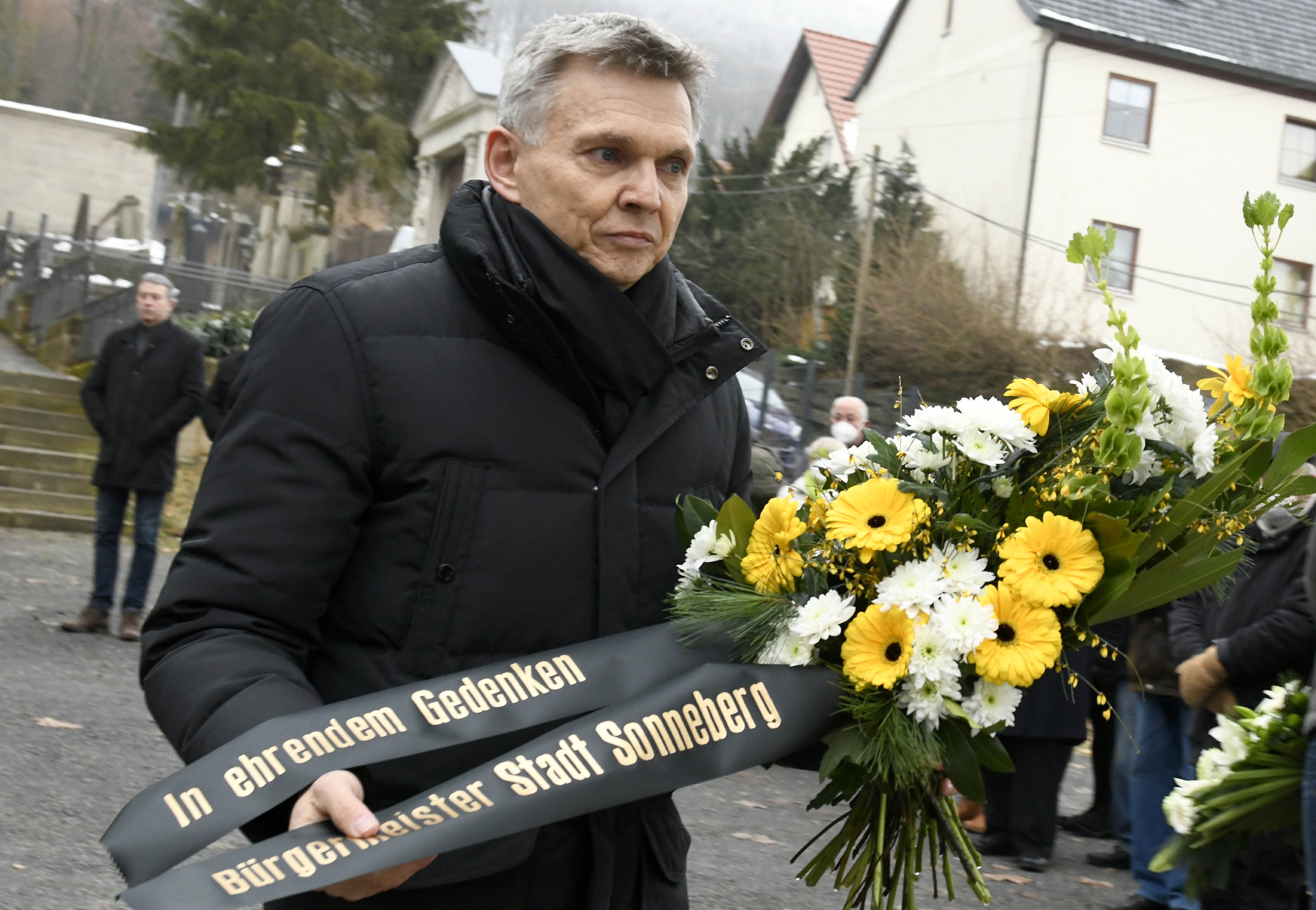 Ein schwarz gekleideter Mann hält einen Blumenestrauß mit Trauerband.