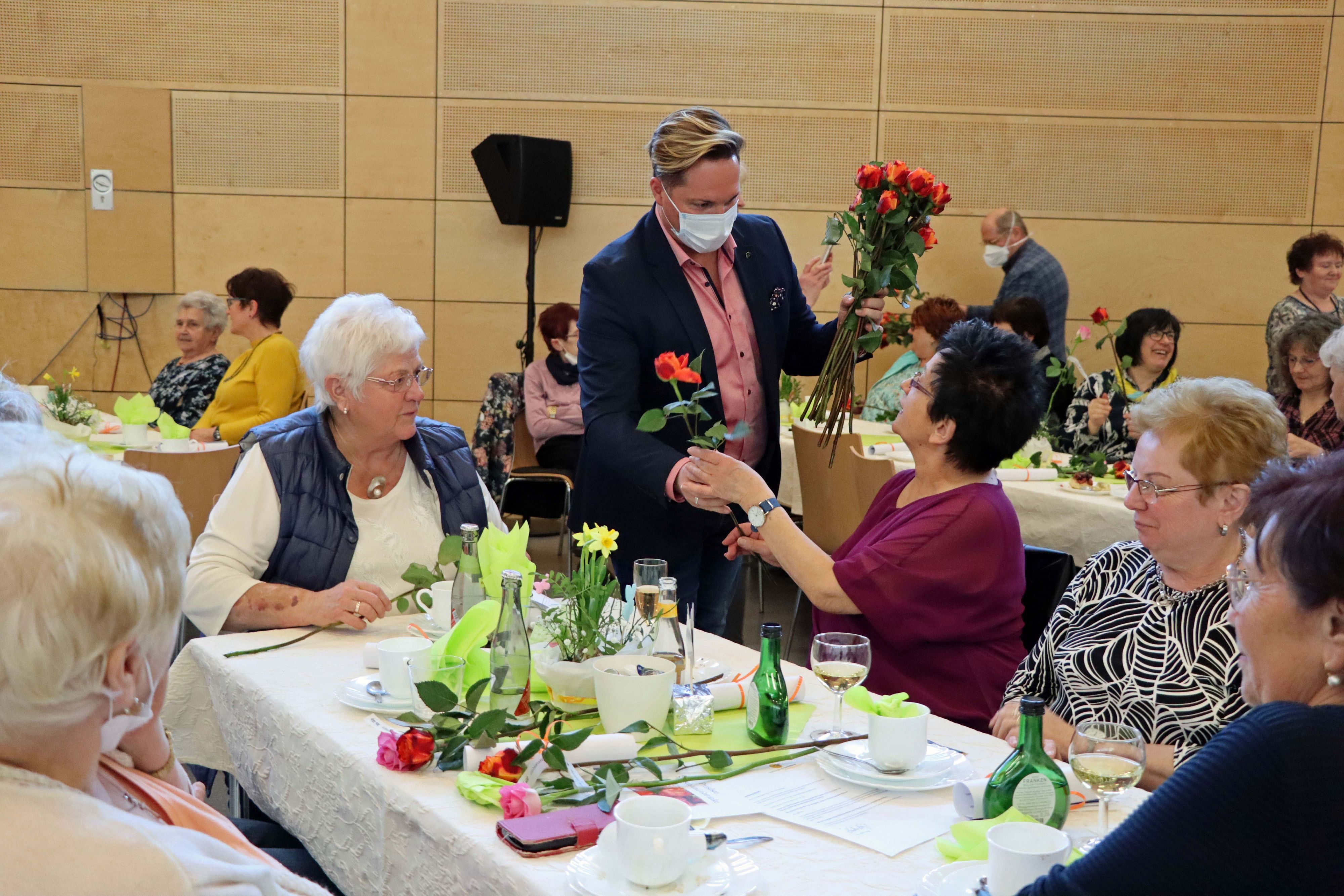 Frauen sitzen an einem Tisch. Ein Mann verteilt Rosen an sie.