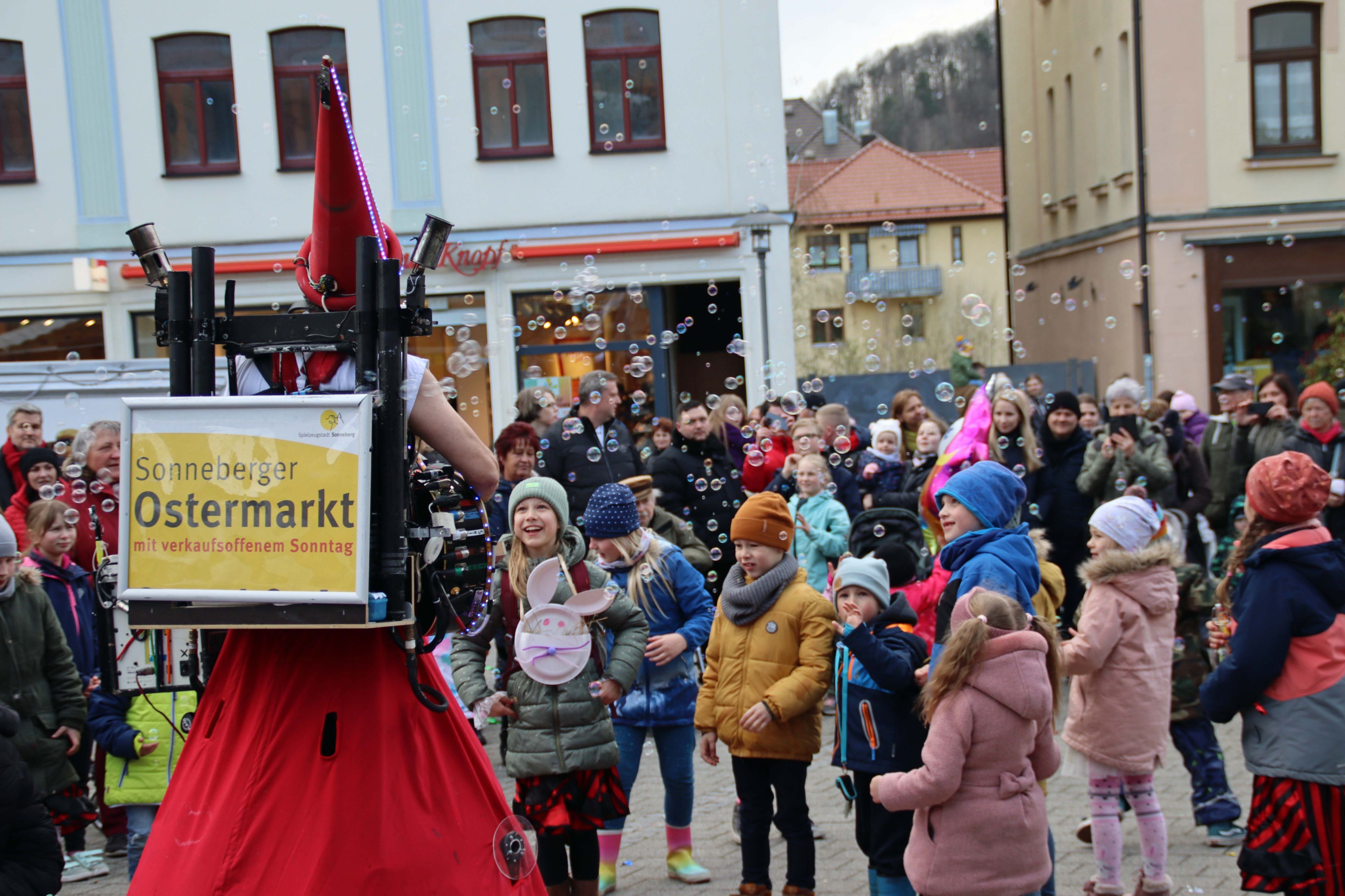 Ein rot kostümierter Mann trägt ein Schild auf dem Rücken mit der Aufschrift: Sonneberger Ostermarkt mit verkaufsoffenem Sonntag. Mit einem Gerät erzeugt er Seifenblasen. Um ihn herum versammeln sich viele Kinder.