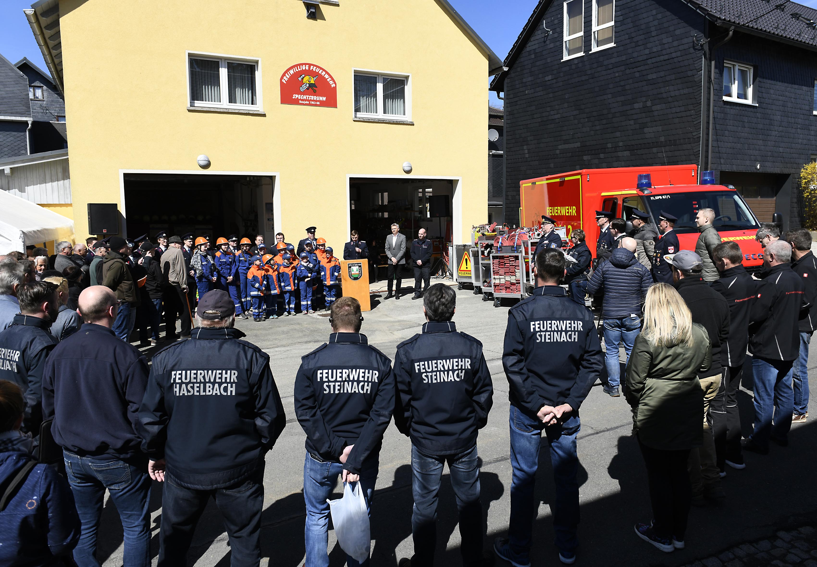 Feuerwehrmitglieder stehen im Kreis vor einem Gebäude. Das Gebäude trägt die Aufschrift: Freiwillige Feuerwehr Spechtsbrunn. Daneben steht ein Feuerwehrfahrzeug.