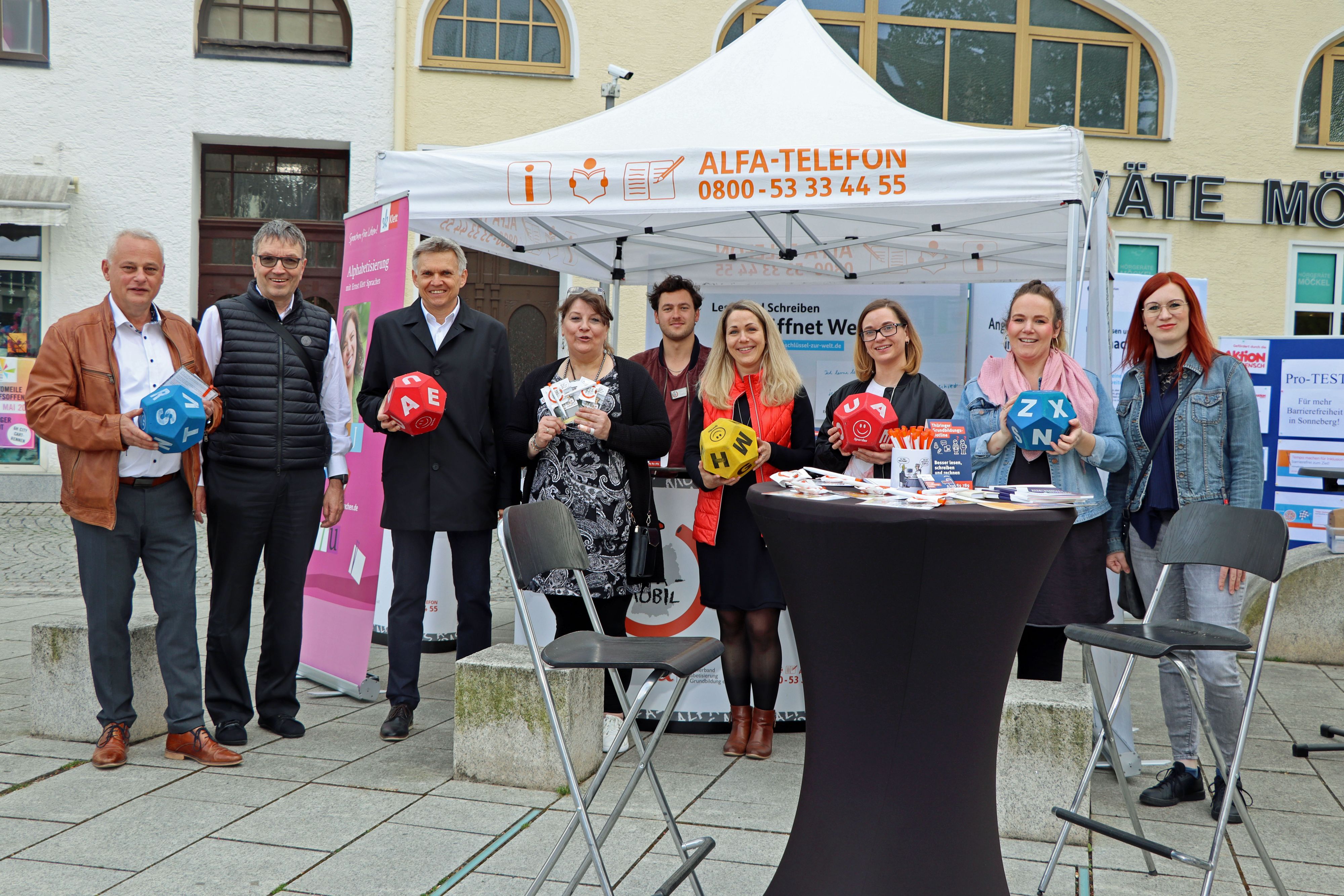 Mehrere Personen stehen in einer Reihe vor einem Faltpavillon. Vor ihnen befindet ein Stehtisch mit Werbematerial darauf.