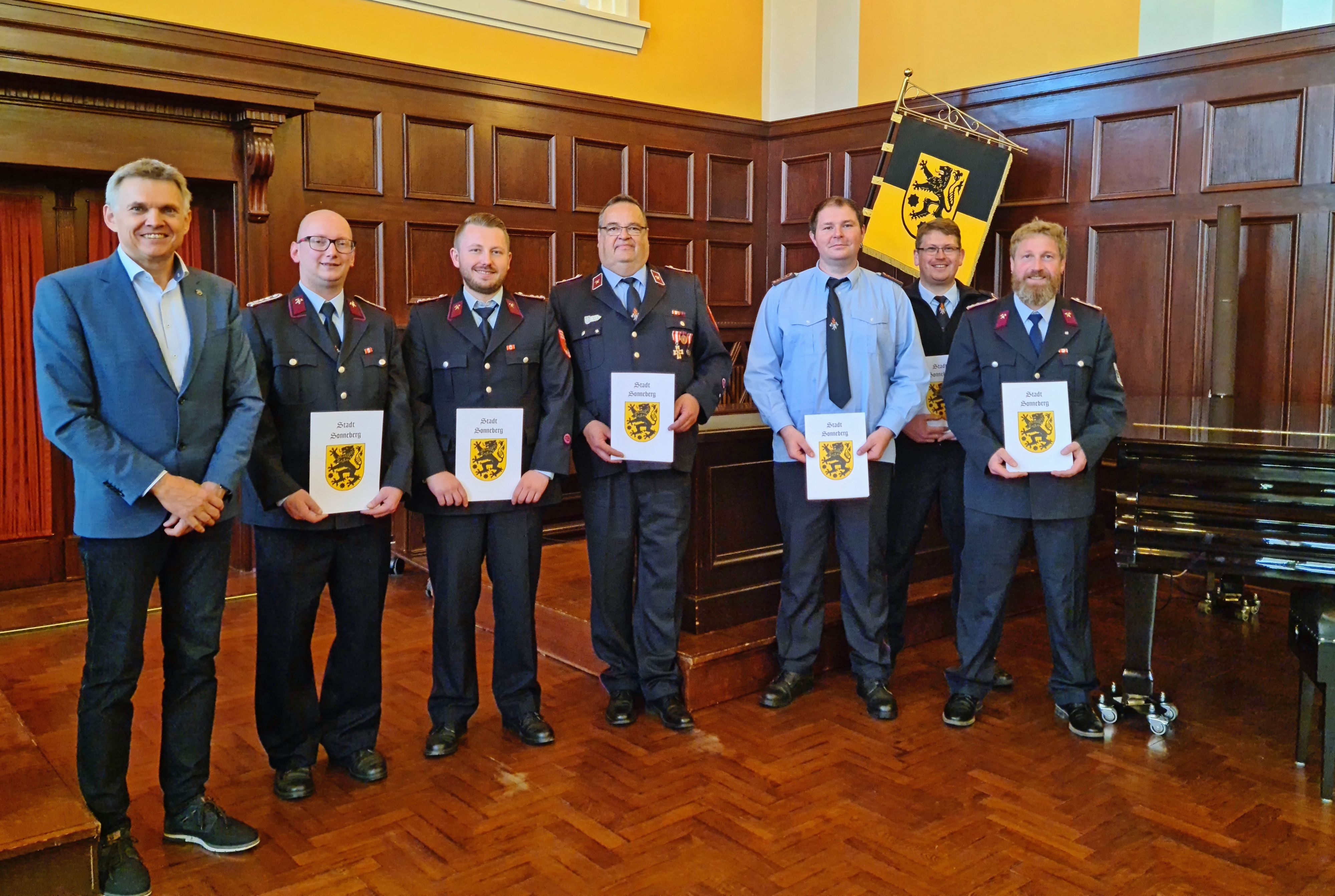 Männer in Feuerwehrunifrom stehen in einer Reihe nebeneinander. Sie halten eine Urkunde in der Hand. Links daneben steht ein Mann im Anzug. Es ist der Bürgermeister der Stadt Sonnberg.