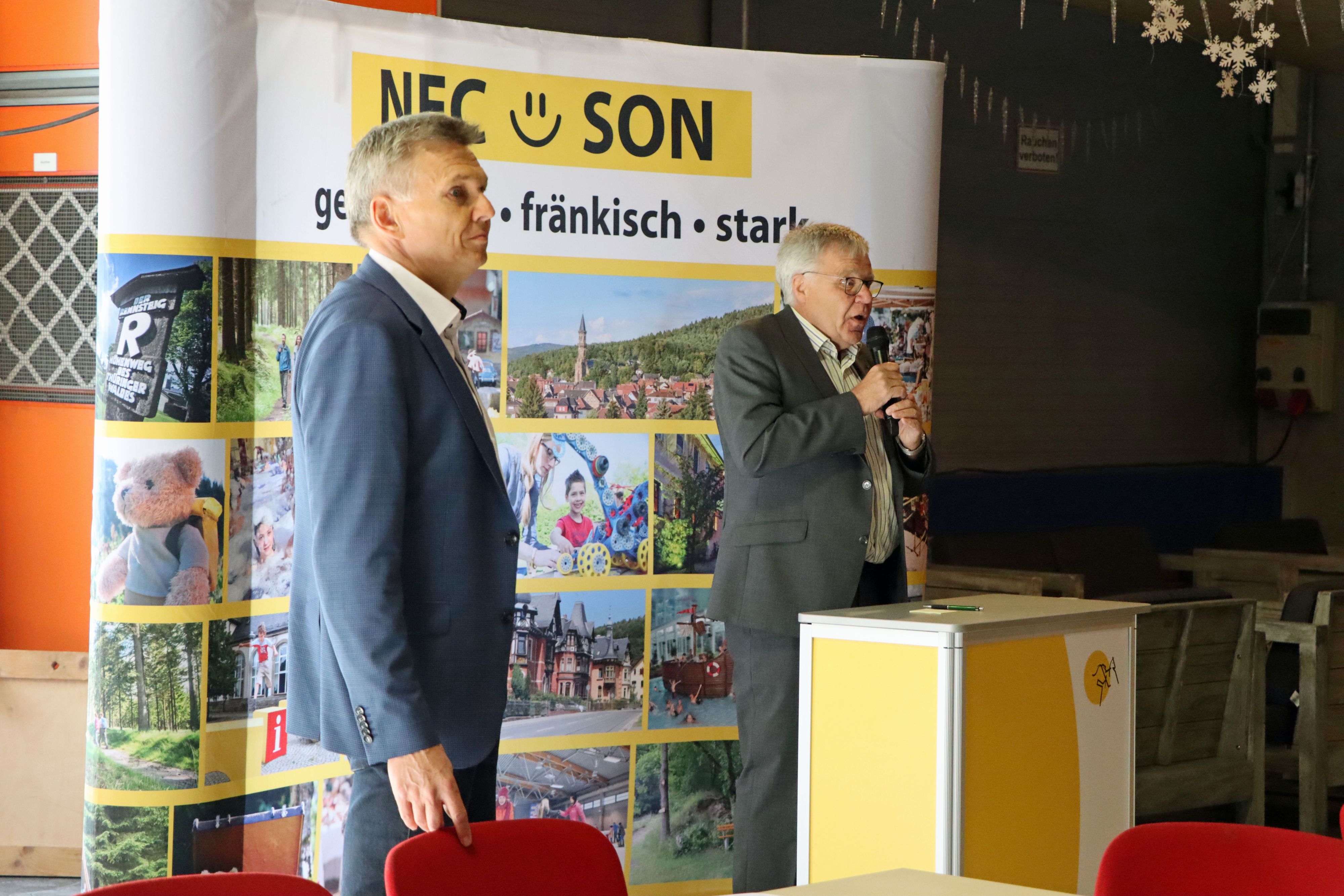 Ein Mann mit Mikrofon steht an einem Rednerpult. Es ist Peter Soyer. Links daneben steht ein weiterer Mann. Es ist der Bürgermeister Dr. Heiko Voigt. Hinter den beiden steht ein Banner mit der Aufschrift: NEC - SON; gemeinsam, fränkisch, stark.