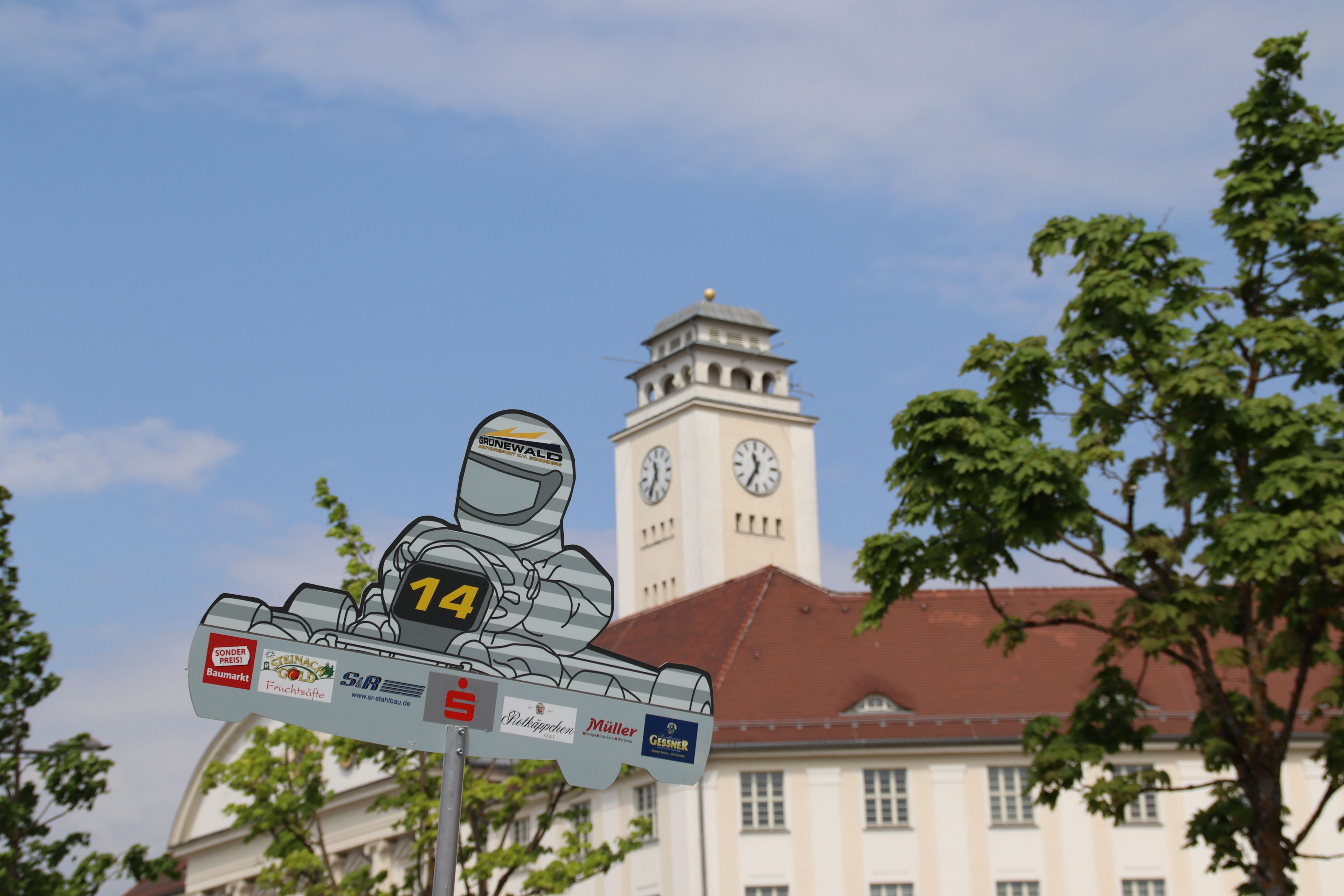 Im Vordergrund ist ein Schild, welches einen Rennfahrer abbildet. Dahinter sieht man das Rathaus der Stadt Sonneberg.