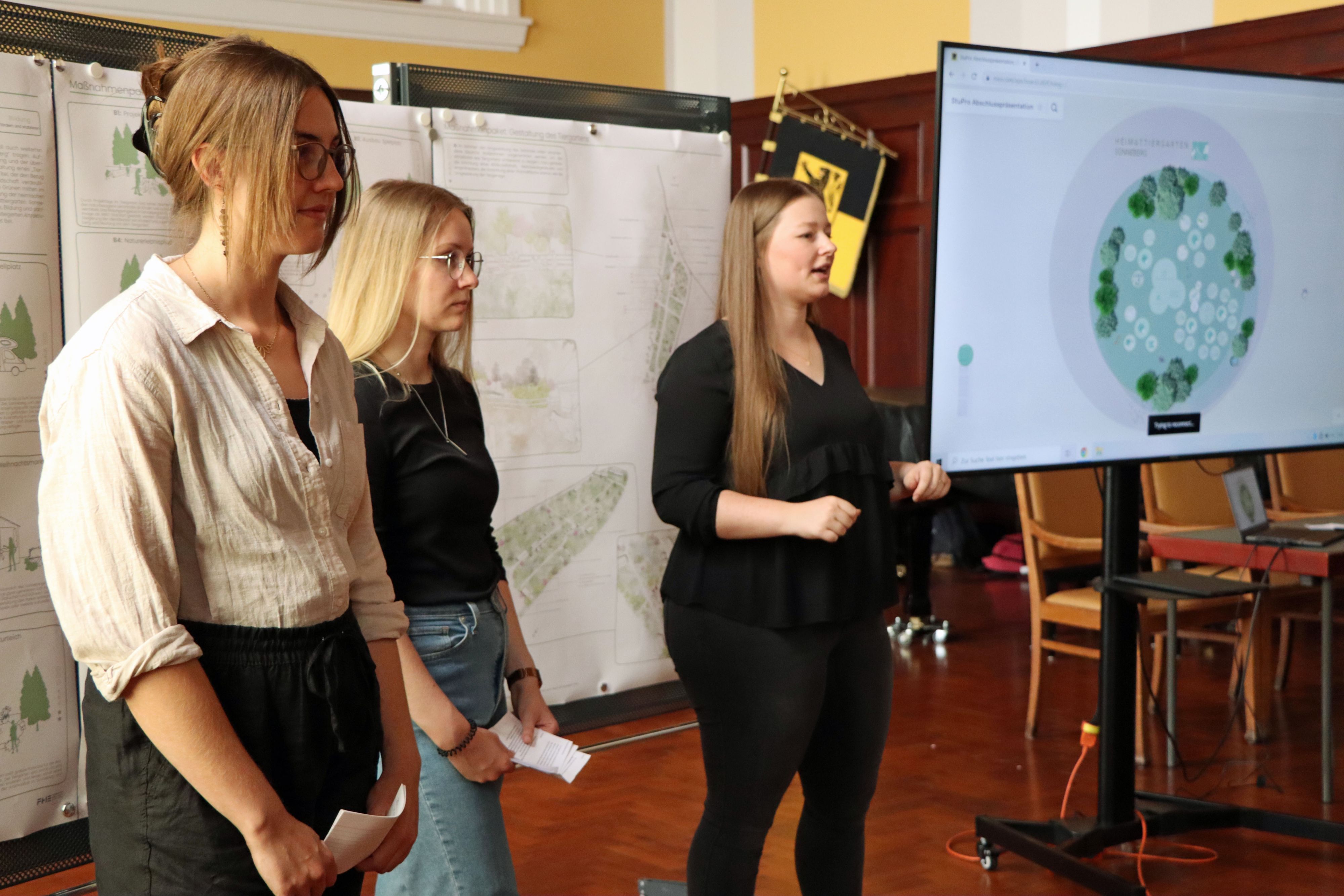 Drei junge Frauen stehen vor Plakaten. Sie halten Karteikarten in den Händen. Rechts daneben steht ein Monitor mit dem Plan einer Grünanlage darauf.