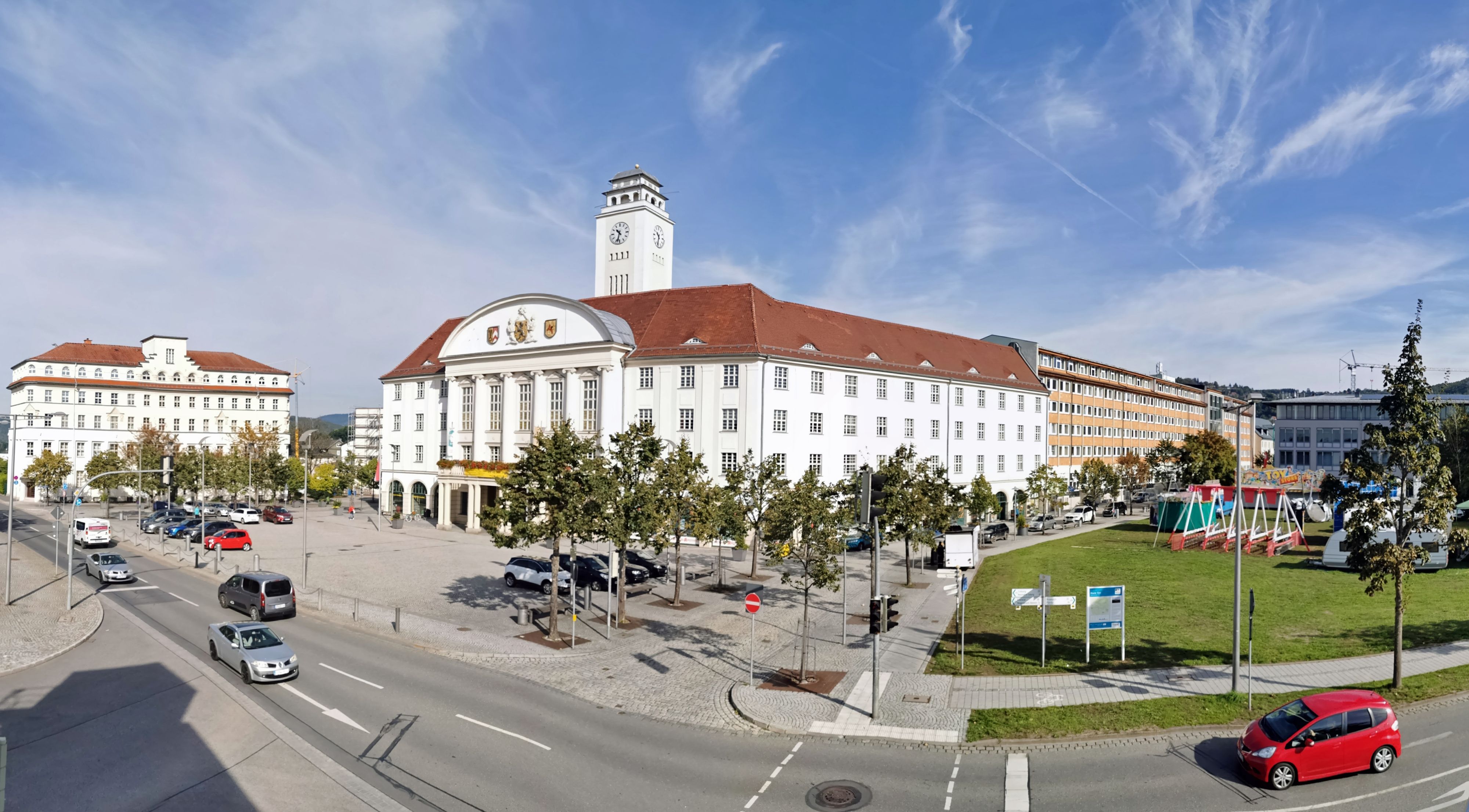 In der Mitte steht ein weißes Gebäude mit einem Turm. Es ist das Rathaus der Stadt Sonneberg. Rechts davon ist eine Wiese. Auf der linken Seite steht das AOK-Gebäude. Die Sonne scheint.