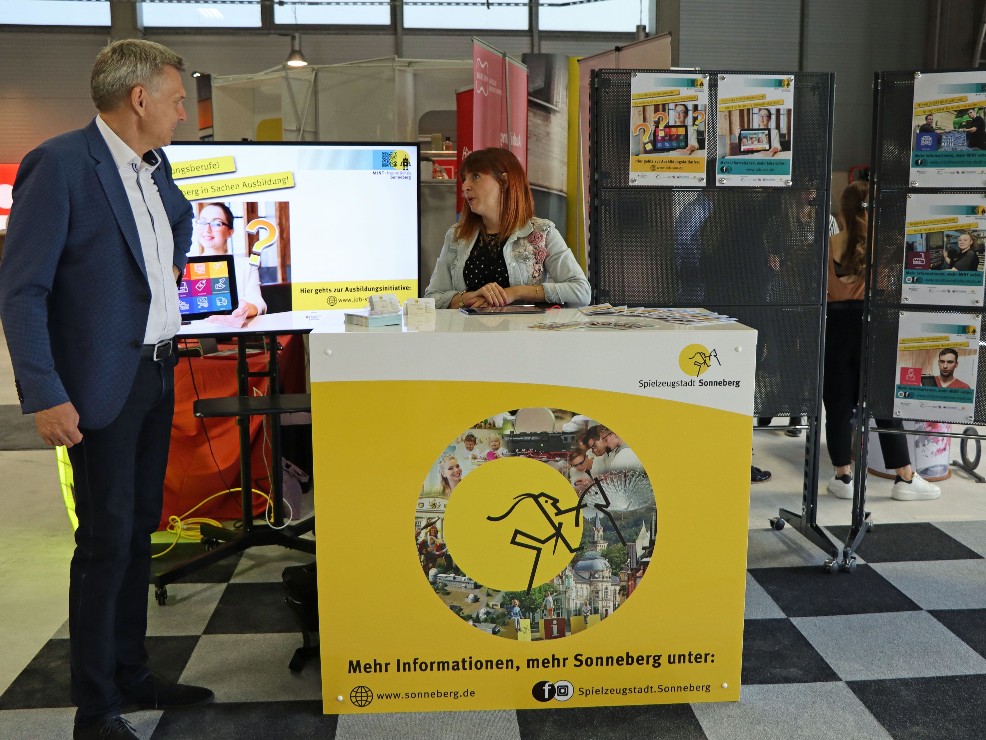 Eine Frau steht hinter einem gelben Messestand. Auf der Standvorderseite ist das Logo der Stadt Sonneberg - das Sonneberger Reiterlein. Daneben steht der Bürgermeister. Im Hintergrund steht ein Bildschirm mit der Aufschrift: Ausbildungsberufe.