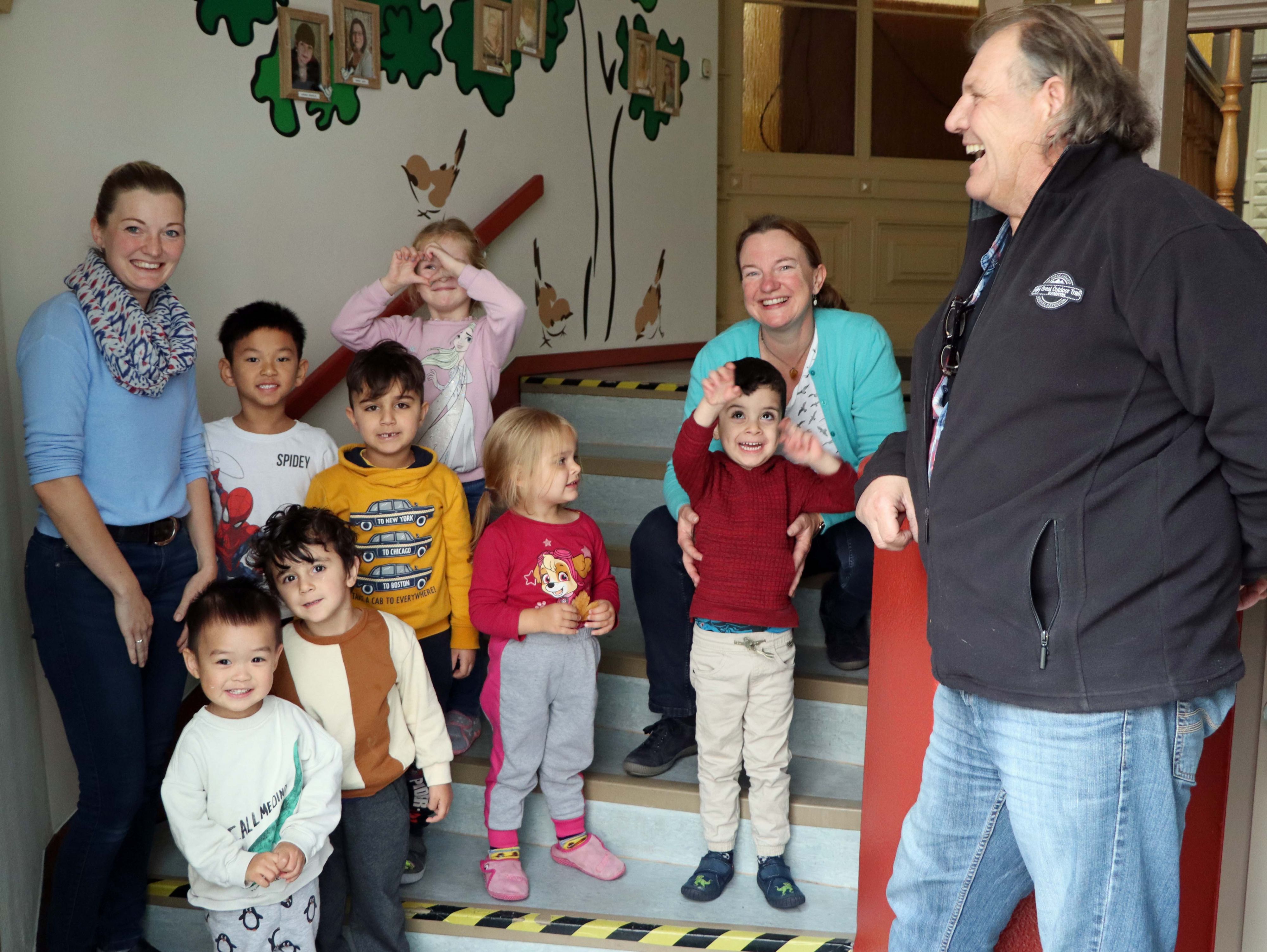 Kinder und Erwachsene stehen in einem Treppenaufgang. Im Hintergrund ist eine mit Bäumen bemalte Wand.