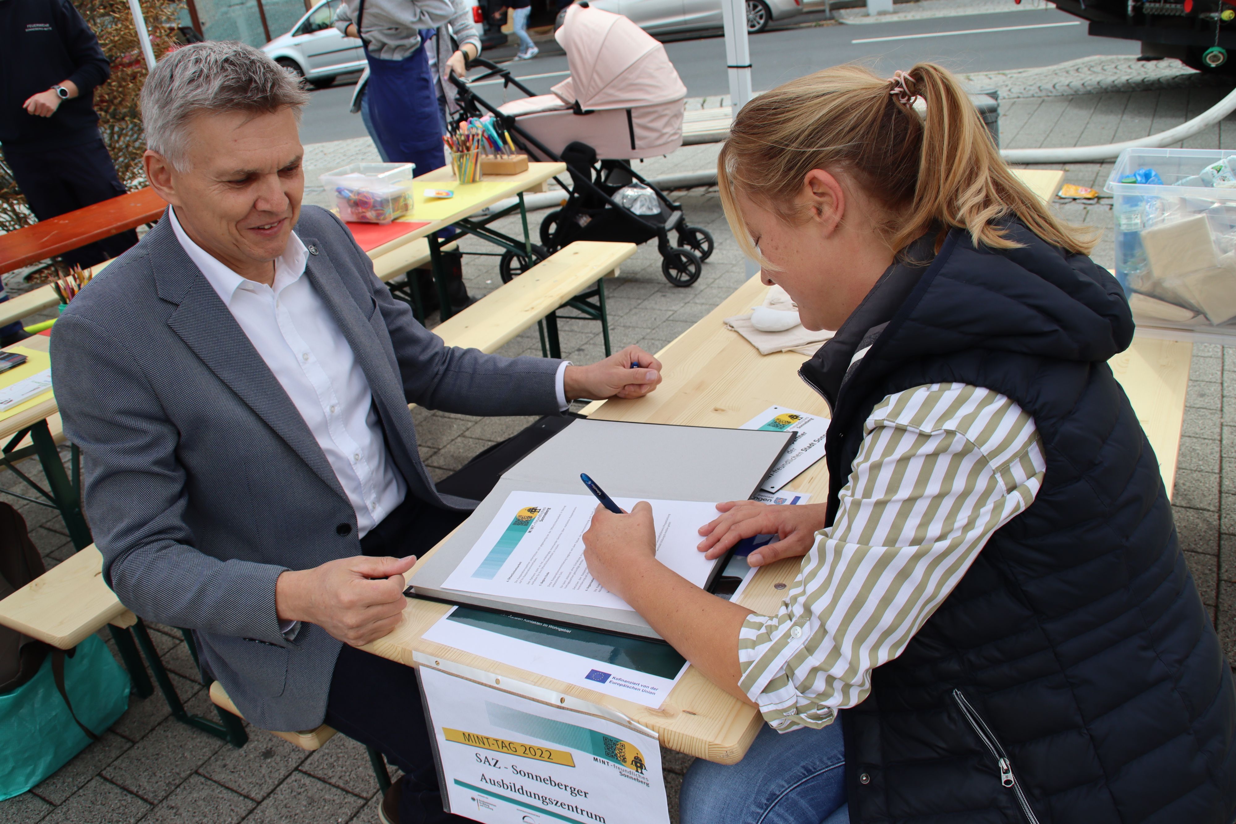 Der Bürgermeister Dr. Heiko Voigt und eine Frau sitzen an einem Tisch. Die Frau unterschreibt Unterlagen.