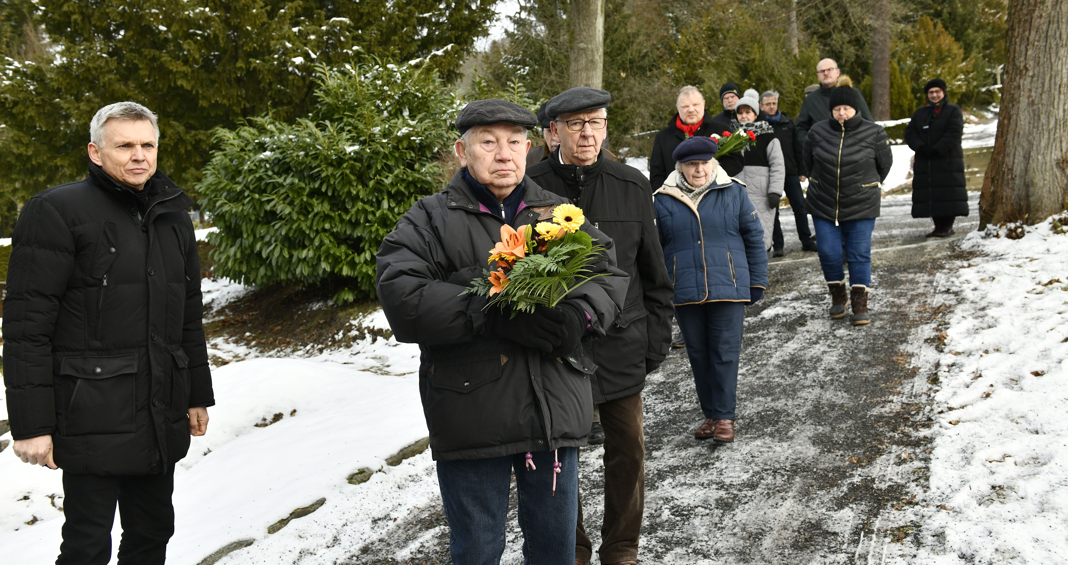 Mehrere schwarzgekleidete Personen bringen Blumen auf einen Friedhof.