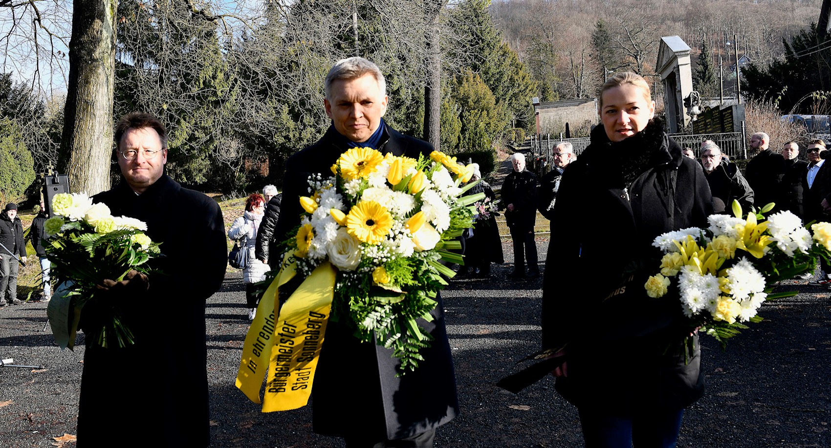 Drei schwarzgekleidete Personen bringen Blumen auf einen Friedhof.