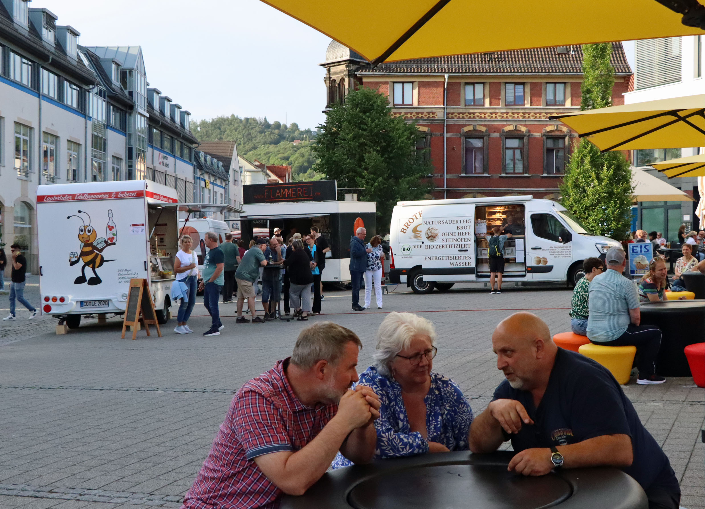 Zwei Männer und eine Frau sitzen an einem Tisch auf einem Marktplatz und unterhalten sich.