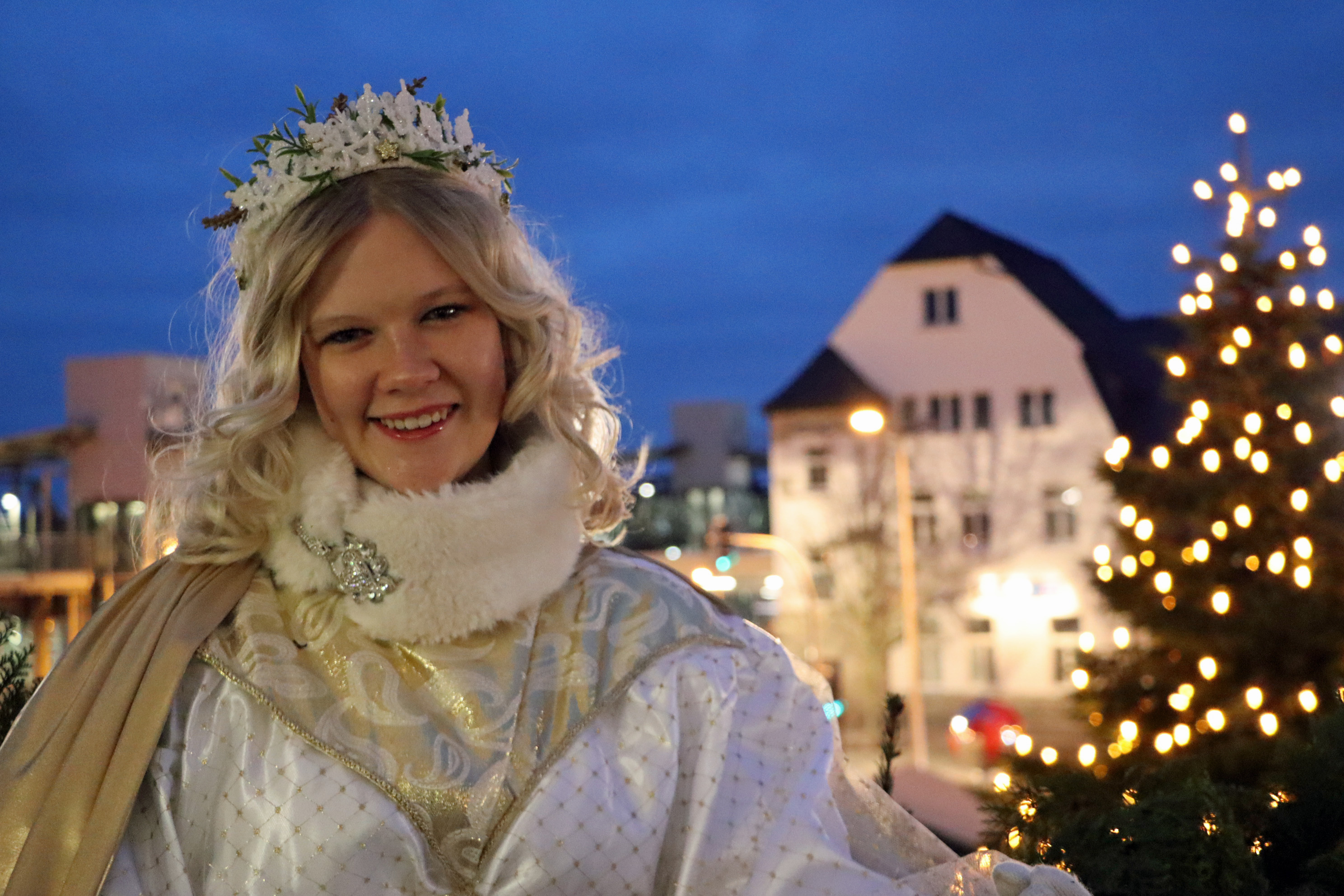 Eine junge Frau mit blonden Locken trägt ein Christkind-Kostüm und befindet sich auf dem Rathausbalkon mit dem Weihnachtsbaum im Hintergrund.